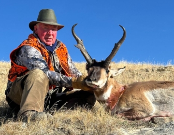 An experienced hunter in an orange hunting vest and patterned camo sits beside a pronghorn antelope on a sloped hill. The bright blue sky provides a stark contrast to the golden grass.